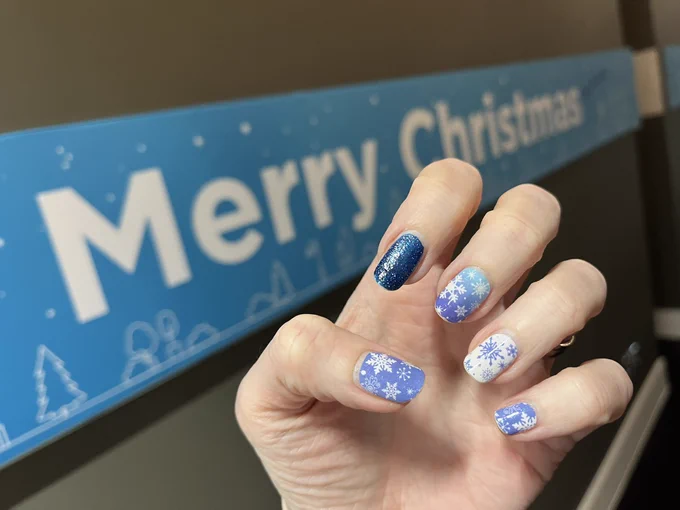 Kate is showing how her nails are decorated with blue glitter and snowflakes, accidentally matching the Merry Christmas banner on the office wall.