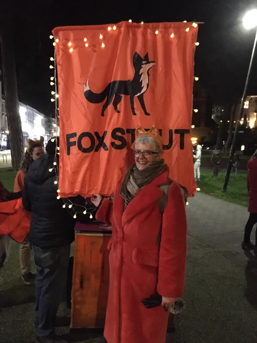 Picture of me, a white women with white, short hair, wearing an orange faux fur coat, standing in front of an orange placard with the illustration of a fox.