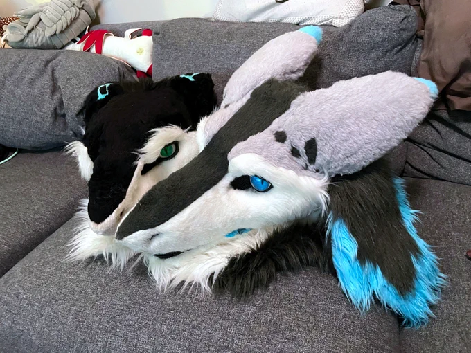 Two Sergal fursuit heads sitting on a sofa, noses pointed towards each other.