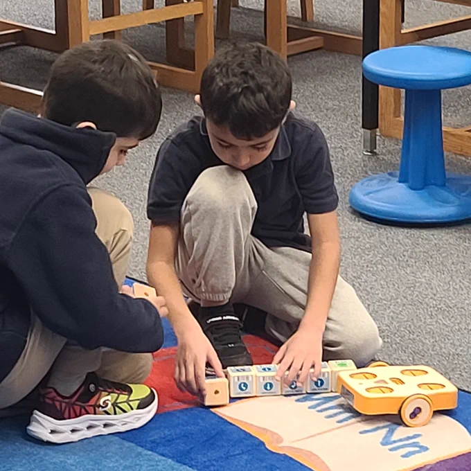Two students kneeling on classroom rug while arranging wooden blocks in front of their Kibo robot 