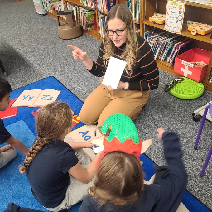 Teacher and students sitting on classroom rug with teacher holding up white card and pointing