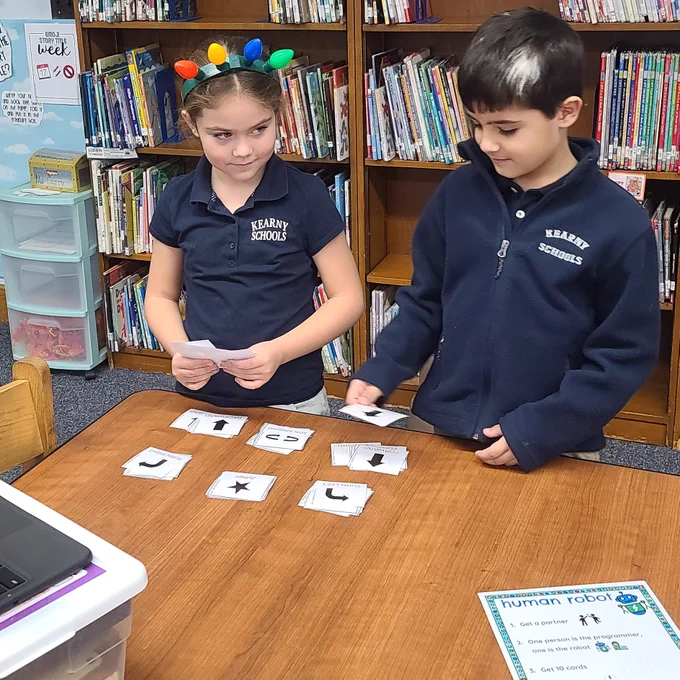 Two students stand at table placing down cards with coding symbols such as arrows and stars