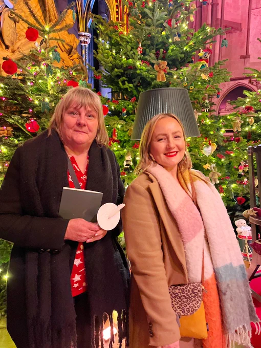 Volunteer Sue and our Chair Megan smile in front of Christmas trees inside the Abbey.