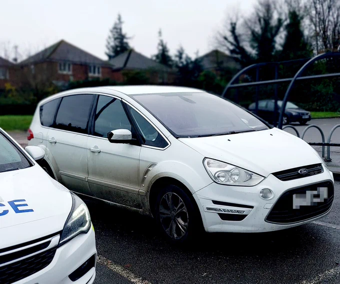 Image shows a large white Ford S-Max parked in a parking bay to the right is a metal framed bicycle storage, and a black car parked in the distance. To the left is the front of a Police car, and behind are some houses and trees. 