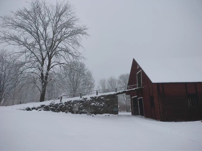 A winter landscape with a red barn and a stone ramp leading up to its second floor. In the background there are some snow clad trees.