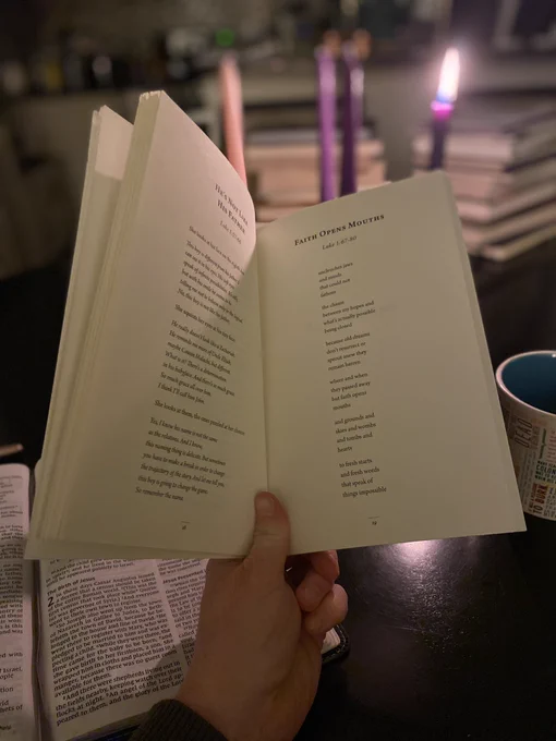 Photo of my hand holding open a book of poetry. In the background is an advent wreath with one candle lit, and a cup of tea