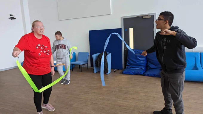 Two people face each other, both holding a gymnastic ribbon. The woman on the left is holding a yellow ribbon and is advising the man on the right how to move his blue ribbon.  The woman on the left is wearing an orange Dance T-Shirt, and the man on the right is wearing a black hoodie.
