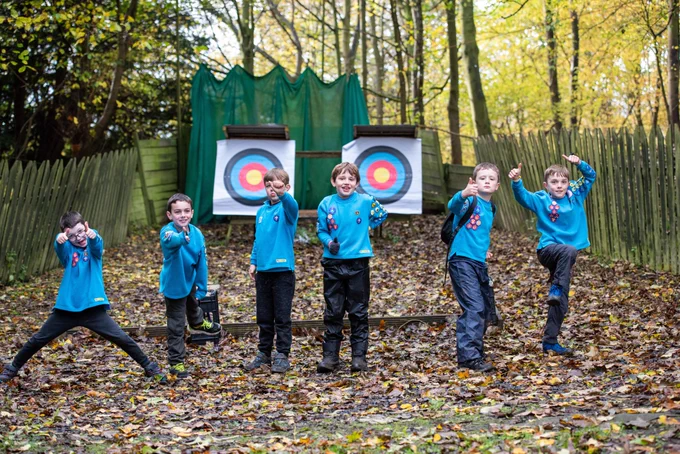 group of 6 beavers posing at the camera and sticking their thumbs up. they are stood outdoors, the ground is covered in leaves and there are archery targets behind them