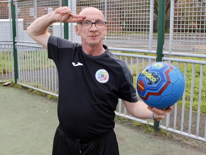 Robert is standing holding a ball and using 2 fingers to salute the camera. 
