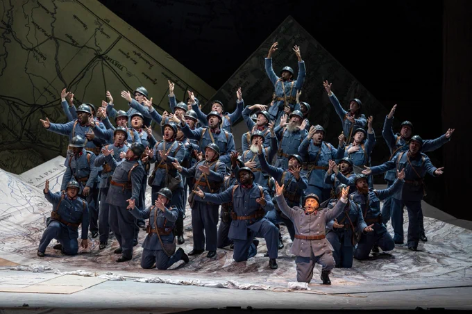 In a scene from Donizetti's THE DAUGHTER OF THE REGIMENT, the soldiers of the 21st regiment are clustered in a group -- mostly standing, a few kneeling, with arms outstretched. They wear blue uniforms with brown belts and metal helmets. They stand on a set of larger-than-life military maps in the shape of mountains.
