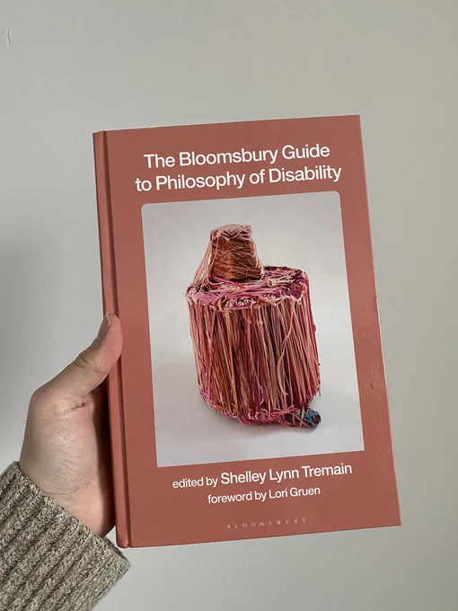 Photo of a hand holding a hardcover book. The book is a salmon colour with an image in the centre. The title of the book “The Bloomsbury Guide to the Philosophy of Disability” is written in white text across the top of the book. The editor, Shelley Lynn Tremain, and the author of the foreword, Lori Gruen, are written in white across the bottom. The image in the centre is a fibre sculpture made of various warm tone strands of yarn, set on a white background.