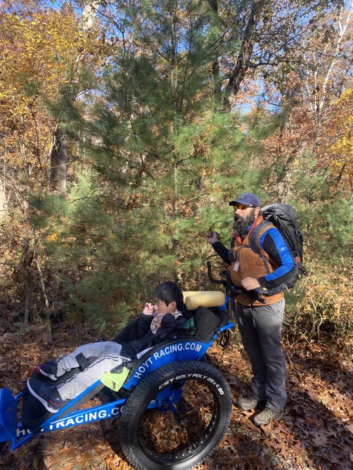 A young man sitting in a Hoyt Racing Chair on a trail surrounded by woods being pushed by Waypoint staff, Rob.