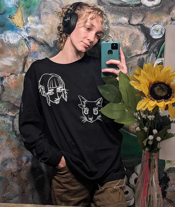 a person with light brown wn curly hair wearing a long sleeve black shirt with a vampire lady and a cat with many eyes on it. the person is taking a mirror selfie in front of a painted wall and a sunflower 