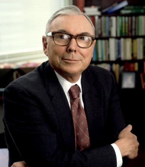 A portrait of Charlie Munger looking directly into the camera, smiling with arms crossed.