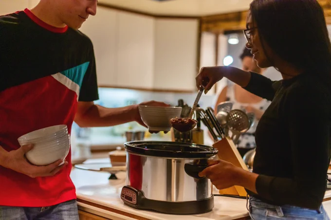 A person serving chili from a slow cooker in a bowl held by another person.