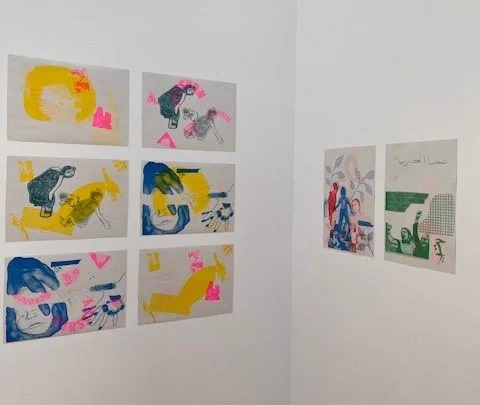 A detail from a corner of an exhibition of riso prints on white walls. The left wall has 6 prints in yellows, blues and neon pink arranged in three rows of two. The right wall has two prints in greens, red and blue side by side.