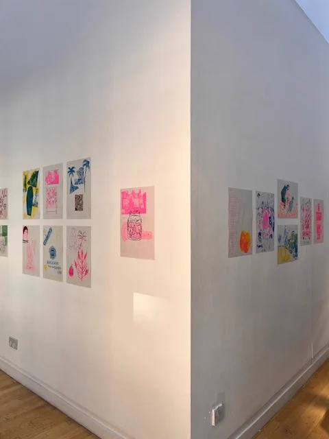A detail from a corner of an exhibition of riso prints on white walls. The left wall has 7 prints arranged in groups and printed in yellows, blues and neon pink arranged in three rows of two. The right wall has six prints in greens, yellow, orange, neon pink and blue in a row.