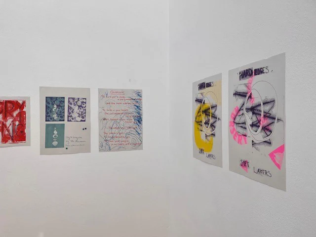 A detail from a corner of an exhibition of riso prints on white walls. The prints are a mix of collage and hand drawn elements in  prints in yellows, purple, red and neon pink. 