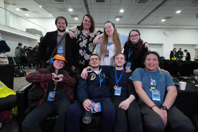 4 people standing behind a couch and 4 people sitting on the coach looking at the camera, with an hotel conference room with rows of chairs in the background.