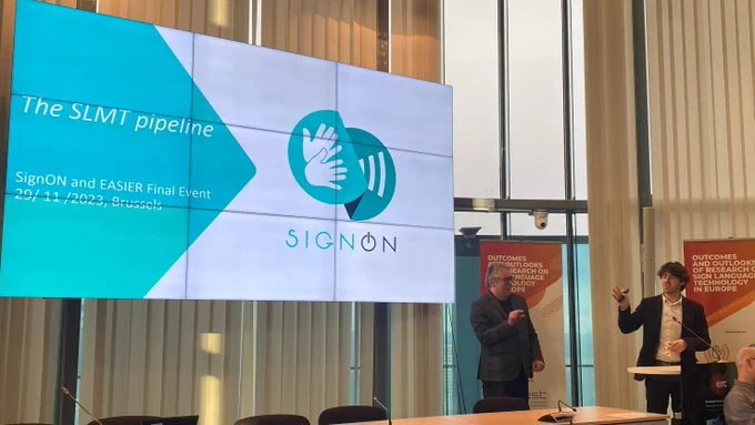 Dimitar Shterionov addresses the audience at the SignON Easier Concertation event. He stands alongside a large screen showing a presentation. It shows the opening slide with the signon logo, titled The SLMT pipeline. He is accompanied by a sign language interpreter. 