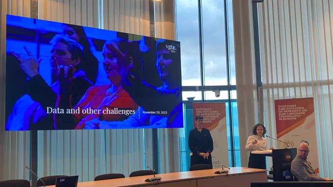Caro Brosens presents at the SignON Easier Concertation event. Caro is accompanied by sign language interpreters, and is standing alongside a large screen with the title slide for her presentation - Data and other challenges. 