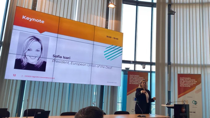 Sofia Isari giving a keynote presentation at the EASIER-SignON Concertation Event, alongside a screen with a slide introducing herself as President of the European Union of the Deaf 