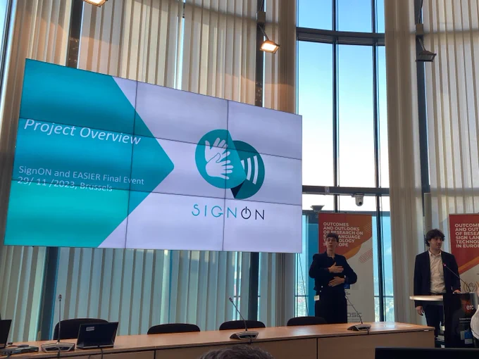Dimitar Shterionov from SignON presenting at today's event, alongside a slide with the signon logo, introducing a presentation on the project overview. He is standing alongside a sign language interpreter. 