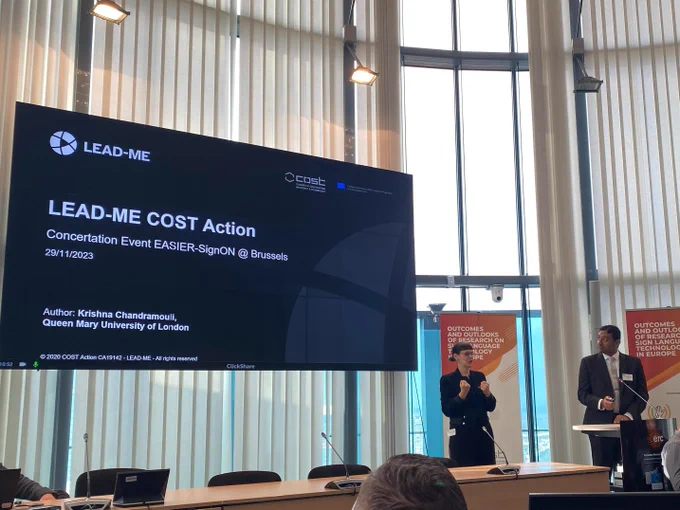 Krishna Chandramouli from LEAD-ME presenting to the audience on the LEAD-ME Cost Action. The slide shows the LeadMe logo, and notes that Chandramouli is based at Queen Mary University of London. He is standing alongside a sign language interpreter. 