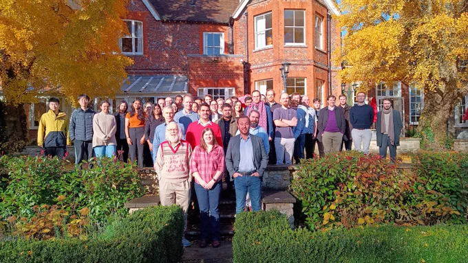 Photograph of a group of people standing arranged on steps in front of a large red brick house, framed on either side with autumnal trees. The leaves on the trees are yellow, and there are red and yellow patches in the low bushes in front of the group. The tiny patch of sky visible is bright blue and some of the people are managing not to wear coats.