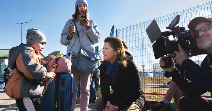 A man is holding a video camera and filming a woman leaning down and talking to a mother and child.