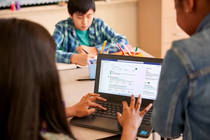 A student sits in a classroom with a laptop open on their desk, a Search Progress assignment is seen on the screen. A teacher looks over their shoulder to give guidance.
