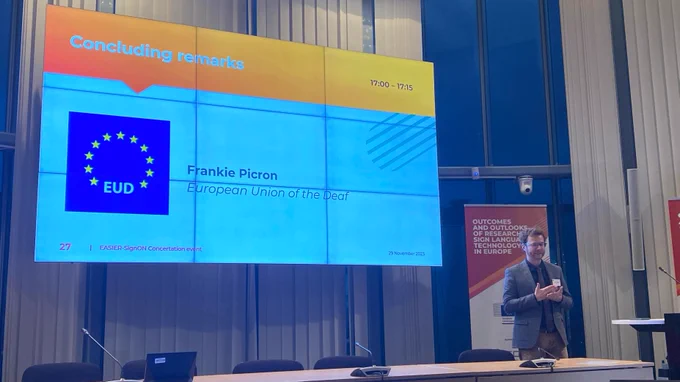Photo of Frankie Picron delivering the concluding remarks at today's event. He is alongside a large screen showing a slide with his name, and his affiliation with European Union of the Deaf. 
