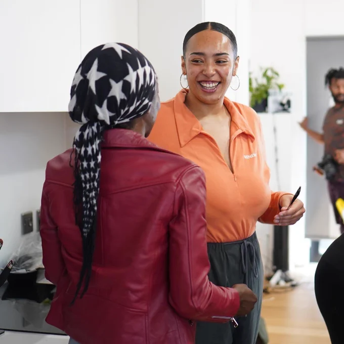 Two people standing at yard and smiling at each other. One has their back to us and wears a red leather jacket with a black scarf with white stars tied round their hair. The other faces us, with their hair tied back and wearing an orange shirt.