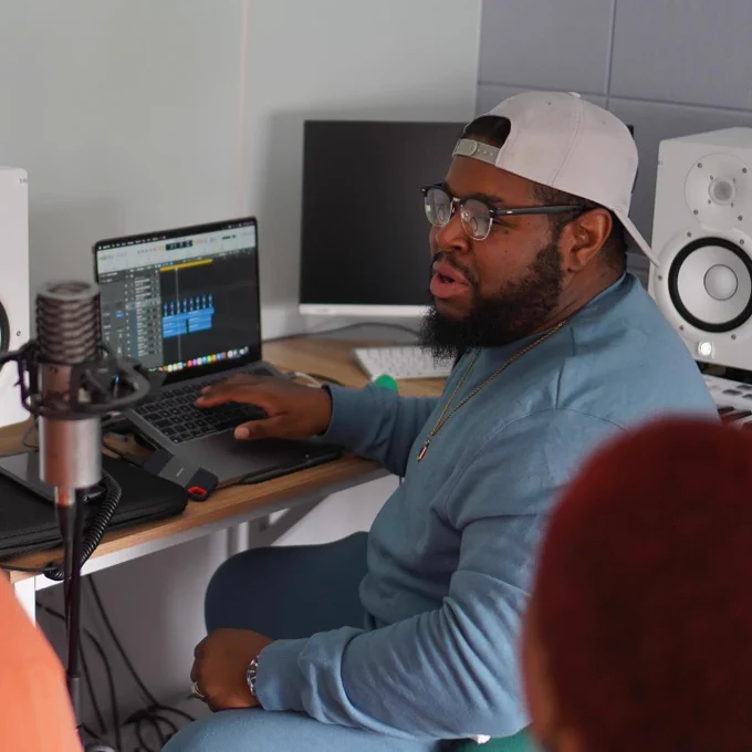 A person in a hat, blue tracksuit, glasses and beard sits in the music studio adjusting levels on a laptop while someone sings into a mic.