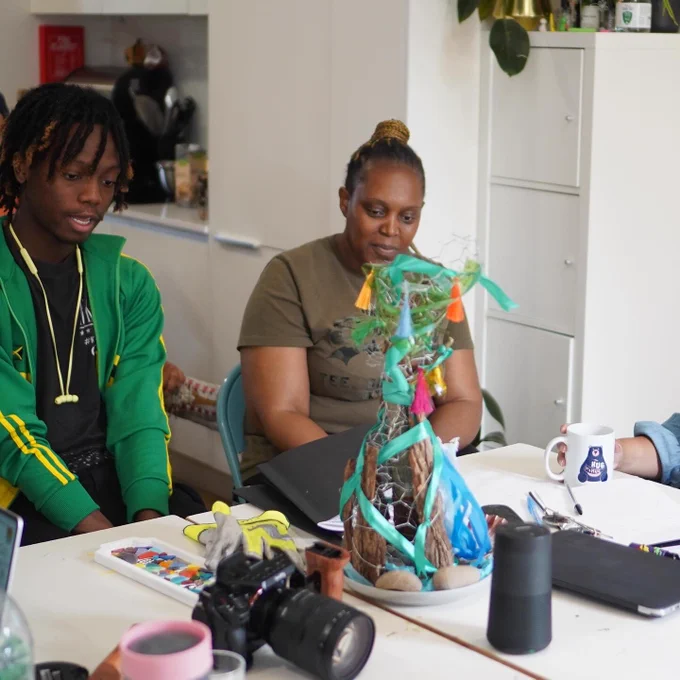 Two people sitting at a table looking at a piece of artwork they have made during art school. It’s a brightly coloured sculpture with bark, metal and ribbons.