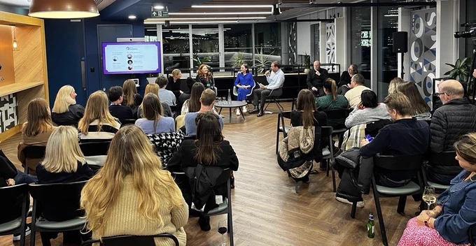 An audience sits in rows in front of three speakers, who are seated at the far end of the room in front of a screen.