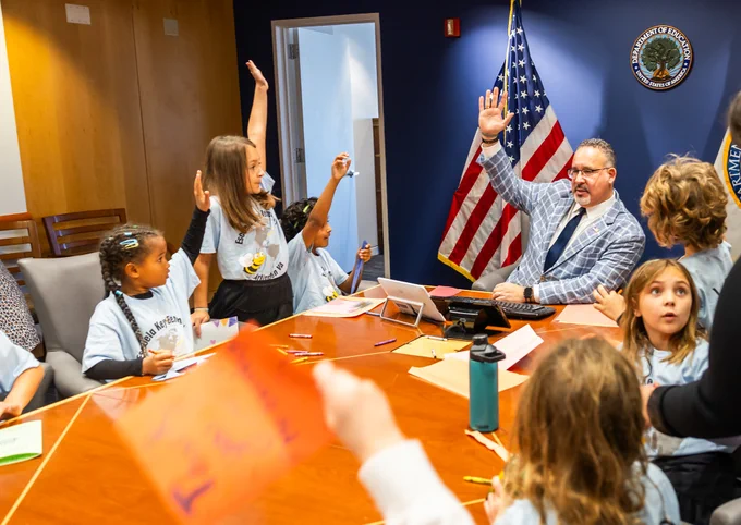Secretary Cardona sitting at desk surrounded by young students raising their hands.