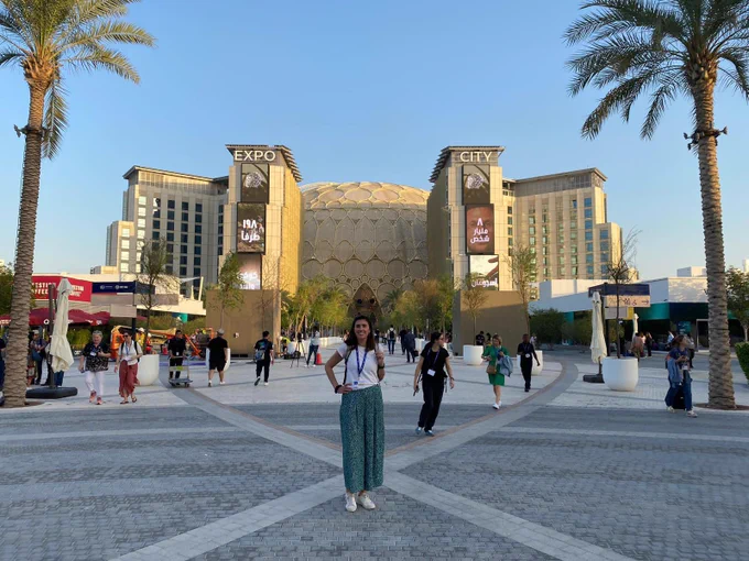 Picture of me standing in front of Expo City, Dubai where the COP28 is being held. It’s a big beige building with a huge dome. The sky is blue and I’m standing in the foreground with my backpack on and a big smile on my face!