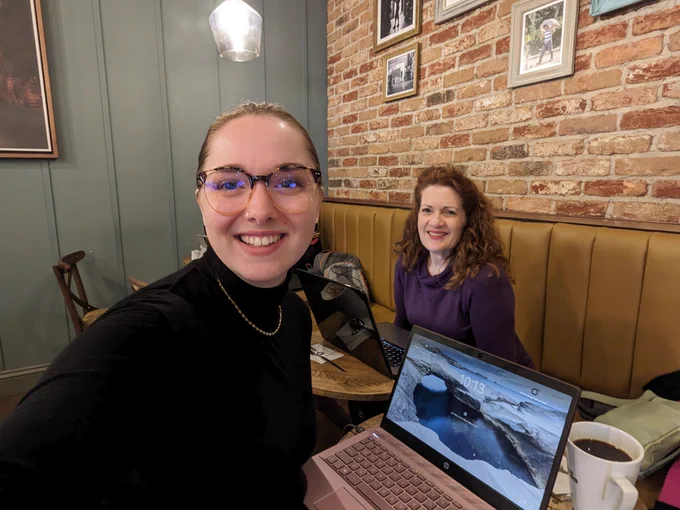 This is a photo of Ruth and Joanne. They are in a coffee shop with their laptops open, smiling at the camera. Ruth is in the foreground and Joanne is slightly further back. 