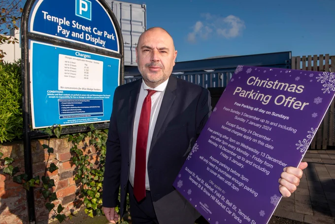 This is a picture taken at Temple St car park and shows a blue sky and a sunny day. Cllr Collingswood is standing next to a car park sign holding a blue board with the Christmas Parking Offer details on it in his left hand.