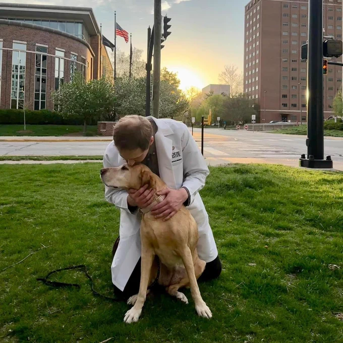 A tender moment is captured between a veterinary student, kneeling on the grass in a white lab coat, and a loving yellow Labrador. He is embracing the dog with a gentle forehead-to-forehead touch, reflecting a bond of trust and care. They are outdoors at dusk, with the warm glow of a setting sun behind the silhouetted buildings and an American flag, illustrating the compassionate spirit of veterinary medicine.