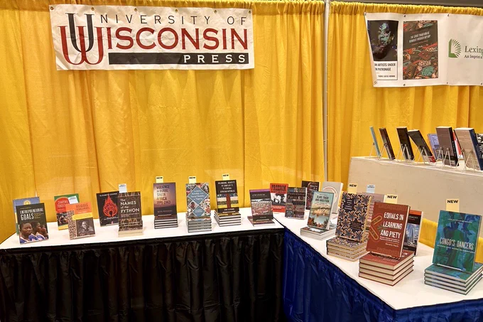 A photo of the UW Press booth at ASA (African Studies Association) conference 2023. In the background, there's a yellow curtain with a banner with the black and red UW Press logo. In the foreground, there's a table with dark blue skirts with lots of recent books on display.