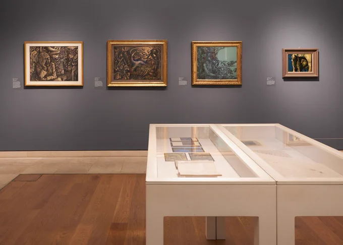 An art gallery with four modernist landscapes on a grey wall, a white wooden vitrine in front of them.