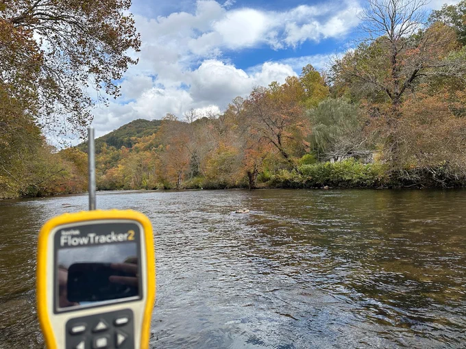 A flowTracker2 instrument with a river, trees, blue sky, and clouds in the background.
