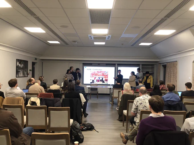 Photograph of a group of seven students standing at the front of a room set up like a small lecture theatre, either side of a projector screen with a slide showing a photo of the Cambridge experimental team. There are lots of people sitting facing them enjoying their presentation.