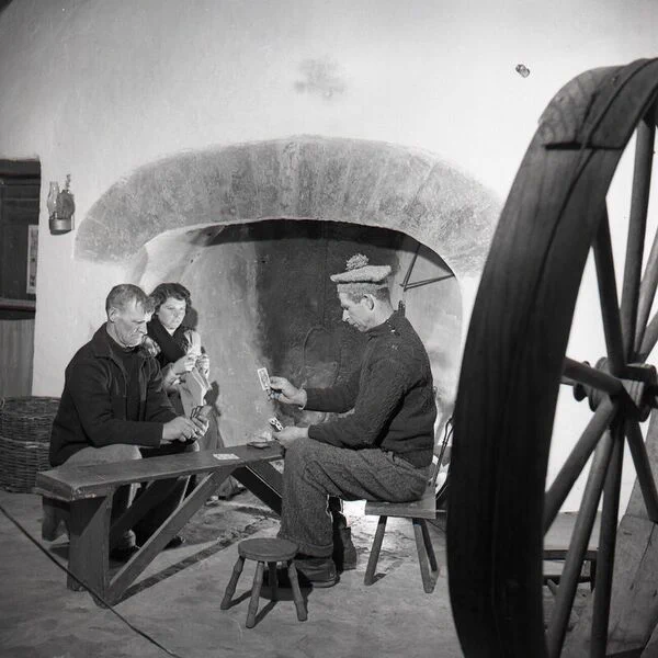 Photograph of two men playing cards on a bench by the fire, with a woman watching while knitting, Aran Islands. The woman is Bridget Johnston. She sits in the alcove of the hearth, in the left midground, behind one of the men. He looks intently at the cards in his hands. The men sit on either side of the bench. The second is Patcheen Faherty. He wears a dark cable knit jumper, dark trousers and a bobble hat. In his left hand he holds a number of cards. In his right hand he holds up a single card, as he looks at the others. Behind them is the hearth, in an alcove. An oil lamp hangs on the wall in the left background. Part of a wheel, possibly a spinning wheel, frames the right of the photo.