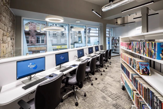 A row of six PCs side-by-side on a long white desk and a white bookshelf with books on the first floor of Paisley library. At the back is the window, through which Paisley High Street can be seen.