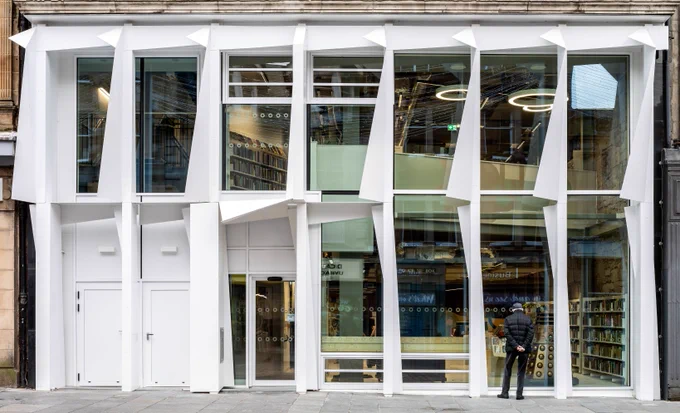 The new modern glass frontage for the bottom two floors of Paisley library. A man in a black jacket and hat is looking through the window from the street.