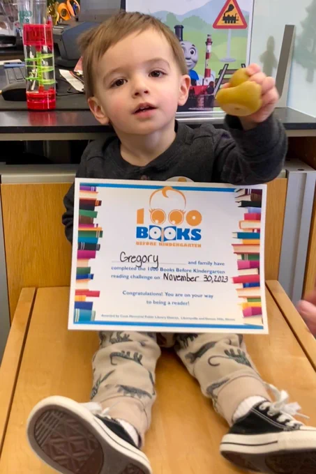 an almost-two-year-old sits on a bench holding a rubber ducky and his 1000 books before kindergarten completion certificate