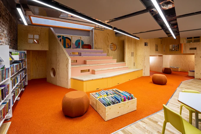 The new children’s library  area with a large wooden play structure and a terraced seated area. There is an orange carpet, bookshelves to the side, and a box of children’s books on the floor.